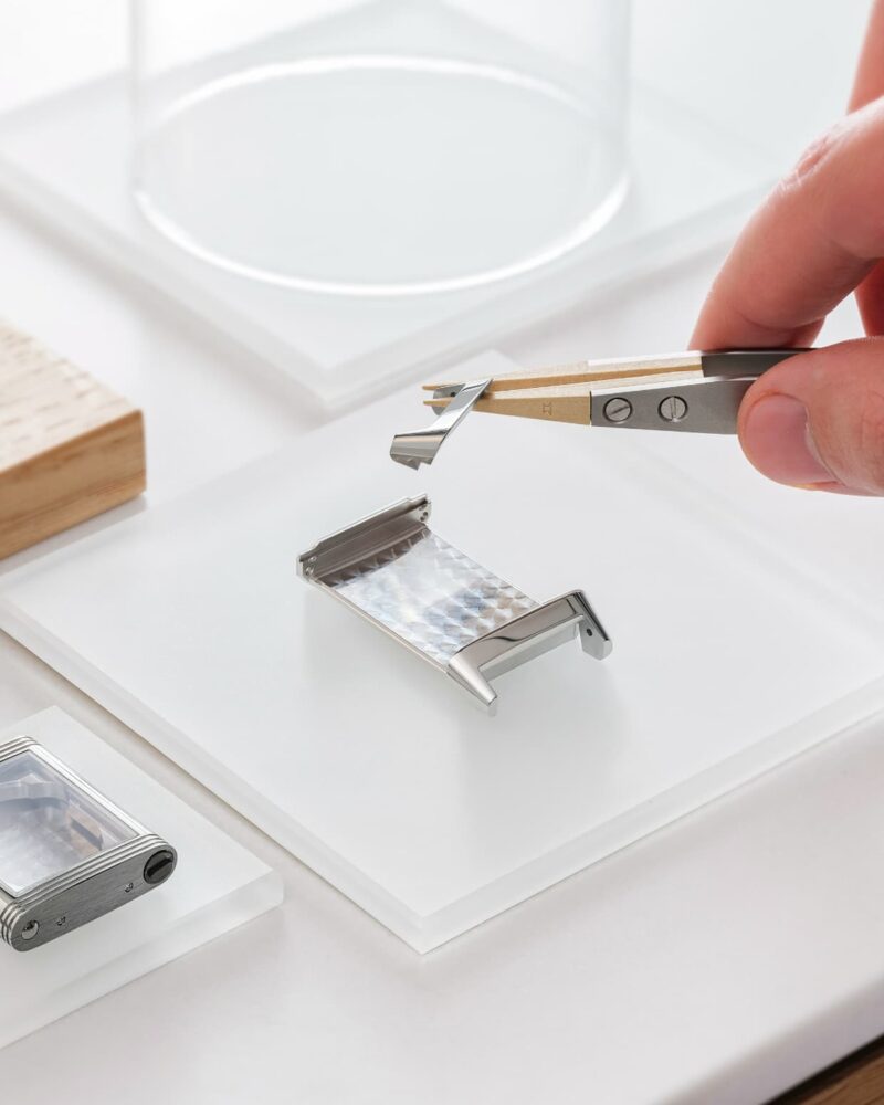 Hand using precision tweezers to hold a watch component above a Jaeger-LeCoultre workbench