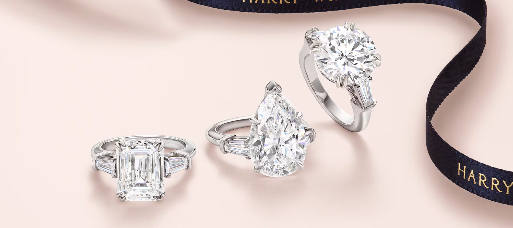 Three different Harry Winston diamond engagement rings arranged on a dark blue silk ribbon with embossed logo