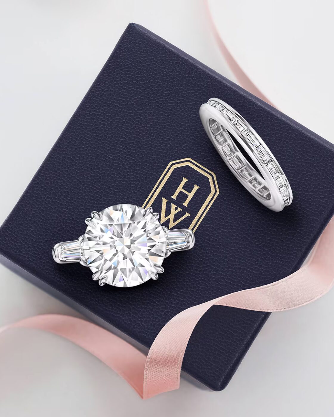 A Harry Winston Classic round brilliant solitaire diamond engagement ring and matching diamond wedding band resting in a navy blue velvet presentation box