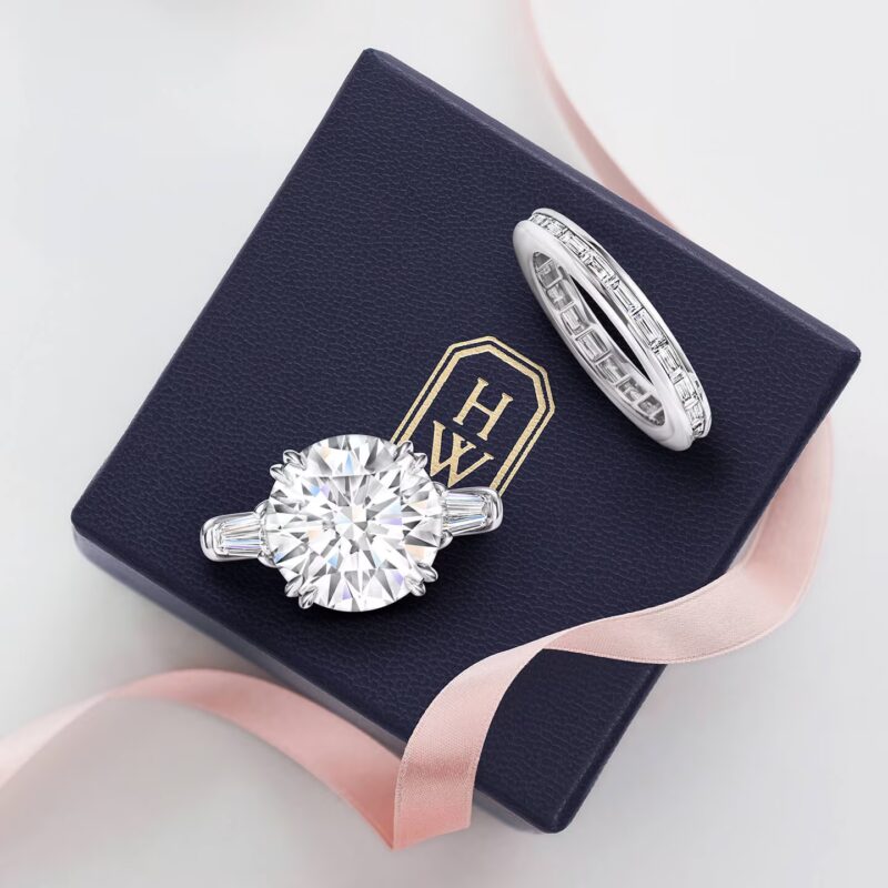 A Harry Winston Classic round brilliant solitaire diamond engagement ring and matching diamond wedding band resting in a navy blue velvet presentation box