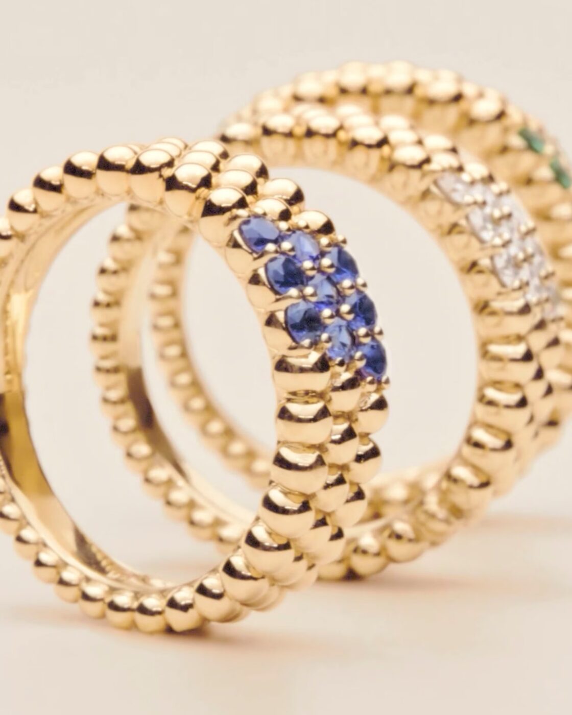 Three Perlée rings standing upright on a beige surface; one with blue sapphires, one with diamonds, and one with green emeralds