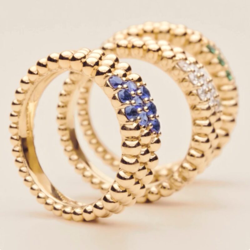 Three Perlée rings standing upright on a beige surface; one with blue sapphires, one with diamonds, and one with green emeralds