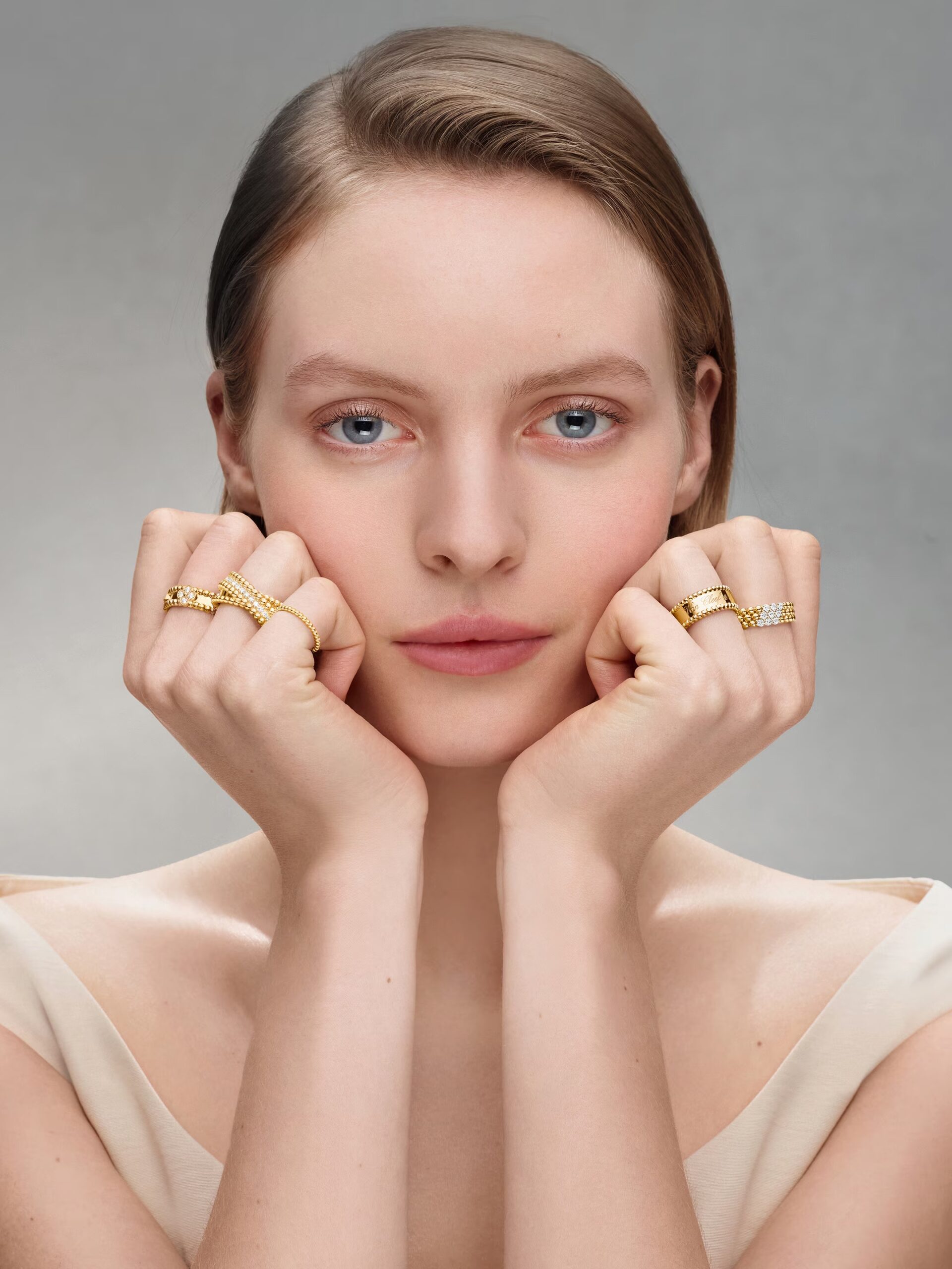 Blonde model looking directly at the camera with her hands under her chin, showcasing multiple Perlée rings (crossover, signature band, pave diamond)