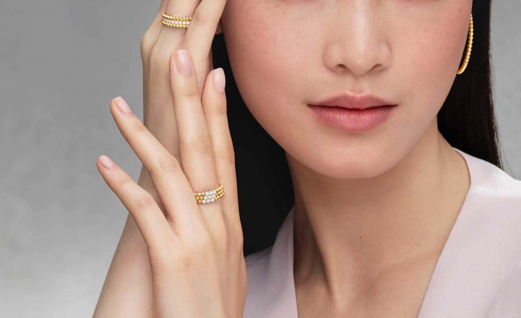 Asian model with hands near her face, wearing yellow gold Perlée rings, a beaded hoop earring, and a thin beaded bracelet