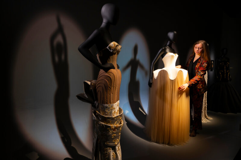Sculptural black-and-gold dress in an exhibition gallery, with a pale gown and a person behind it.