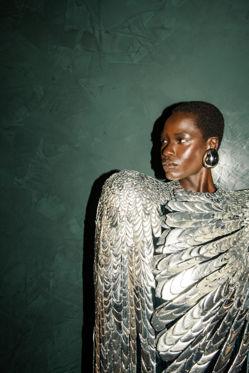 Awar Odhiang wearing a large circular earring beside a broad sculptural pale shoulder and layered couture surface.