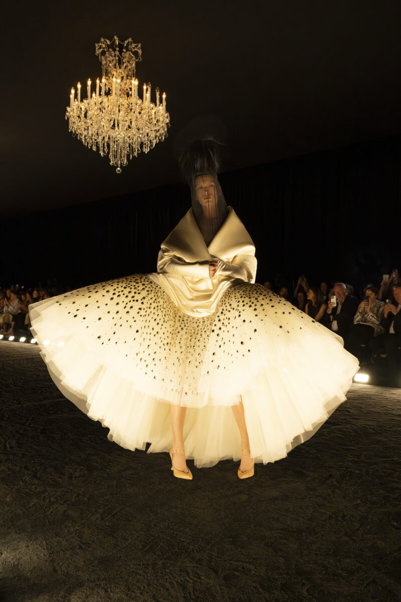 Runway model wearing pale couture with a broad skirt dotted with dark marks and fabric rising around the head.