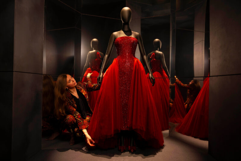 Person crouching beside a red dress displayed on a mannequin in the Schiaparelli exhibition.