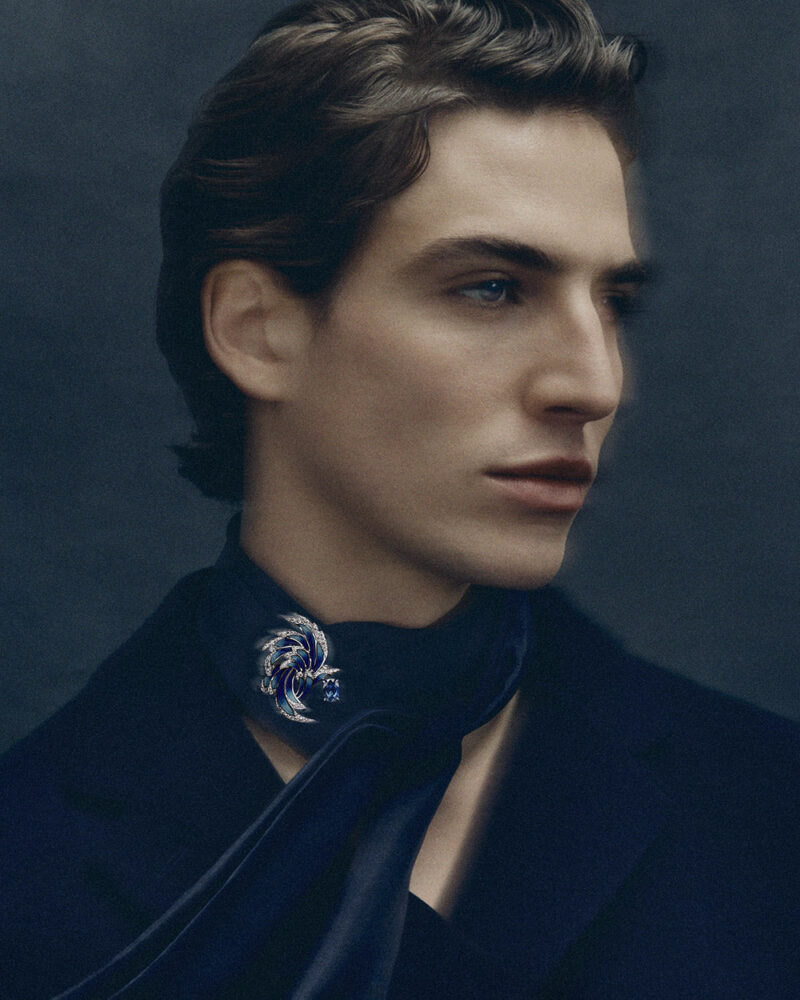 Man in dark clothing wearing a curved blue and bright stone-set brooch near his collar.