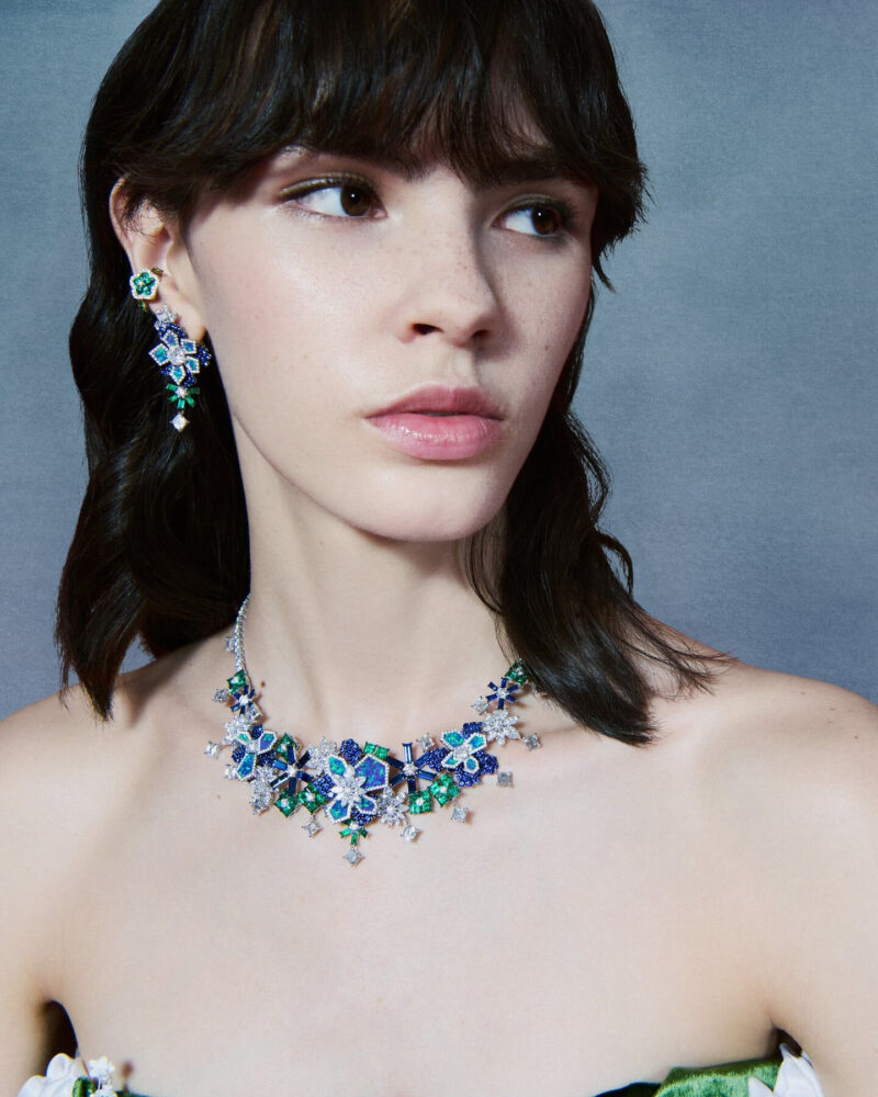 Woman wearing a necklace and earrings formed from blue and green flower-like clusters above a green and white dress.