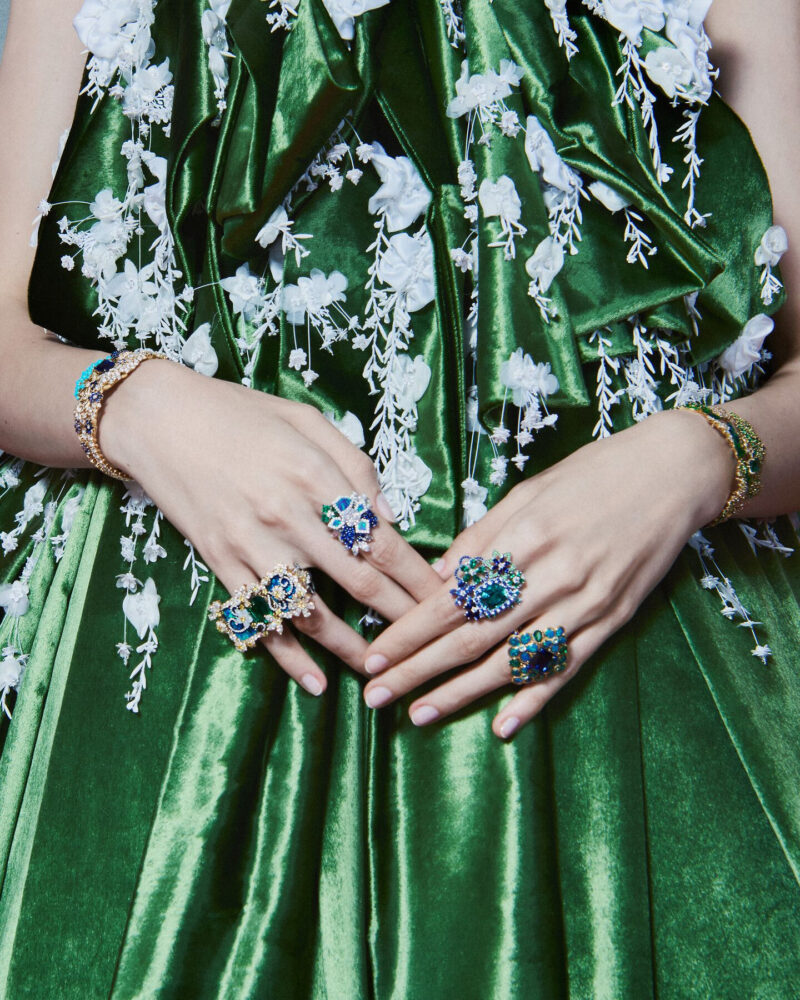 Hands wearing large blue-centred floral rings resting against a green dress with pale floral details.