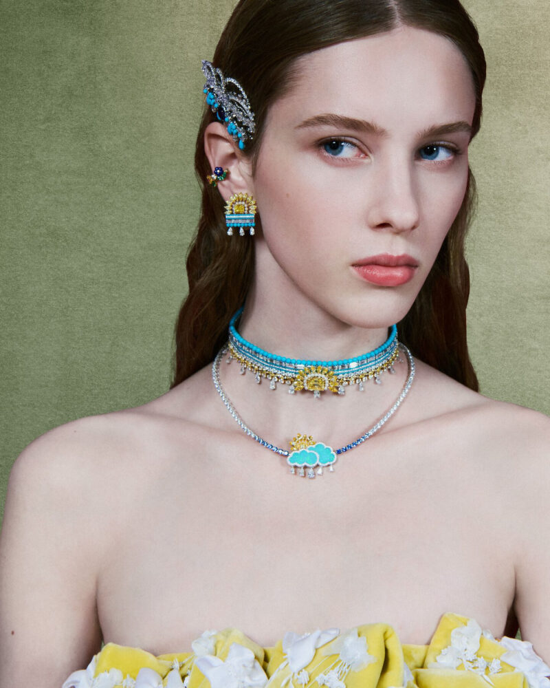 Woman wearing layered blue and yellow necklaces, a cloud-shaped pendant and an ornament beside her hair.