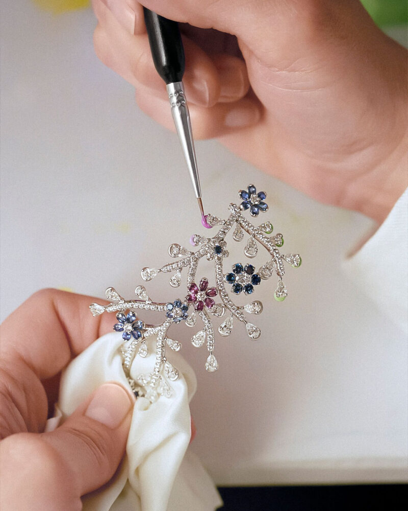 Hands holding a delicate branching jewel while a small brush approaches a pink element.