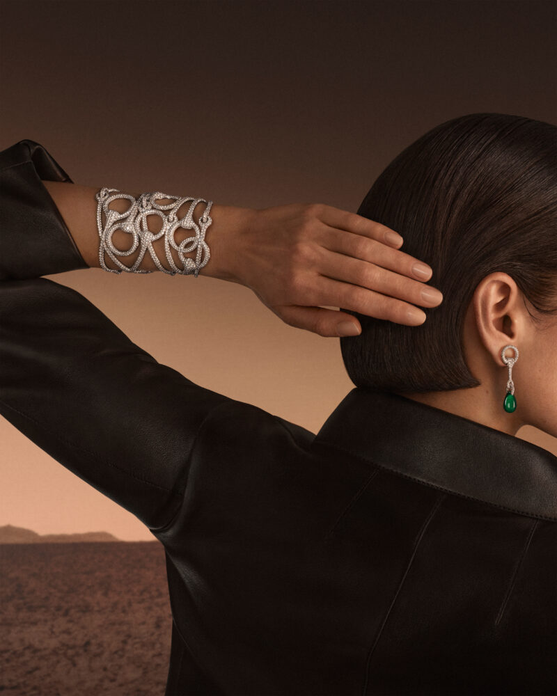 Woman with a hand behind her head wearing a bright open-loop cuff and a long earring beside a raised black collar.