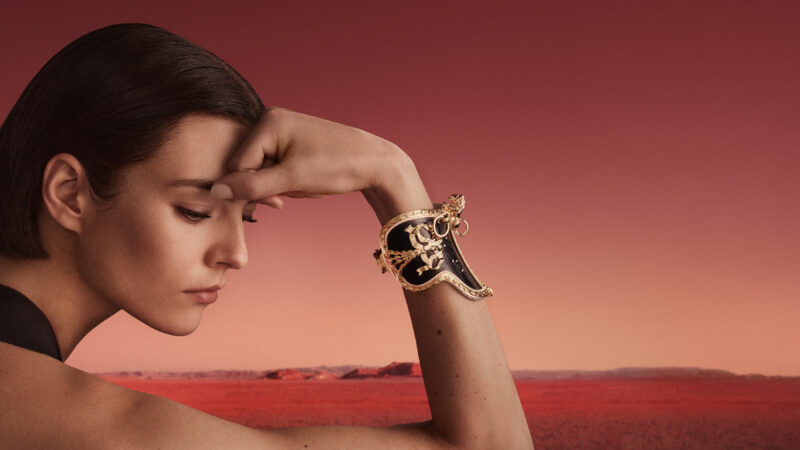 Woman bending towards her raised hand, a dark and gold-toned Sellette bracelet at her wrist against a red landscape.