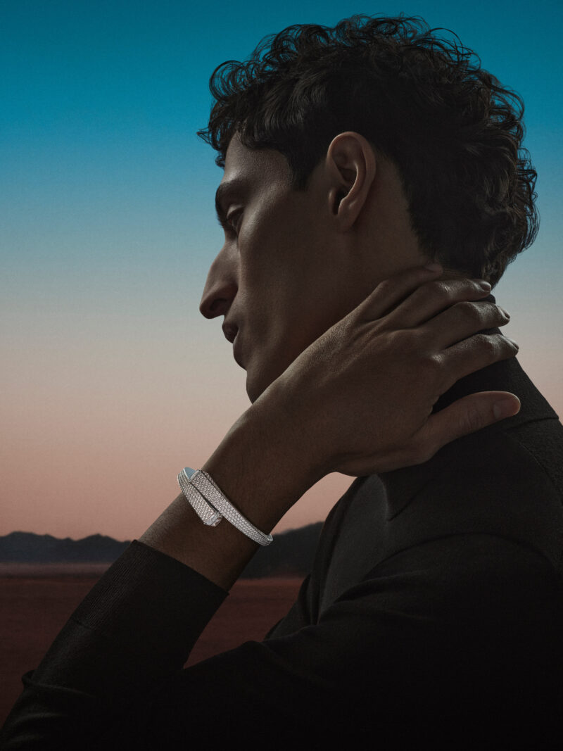 Man holding a hand near his face, a narrow bright bracelet between the wrist and a dark sleeve.