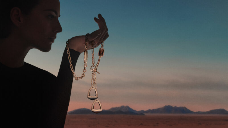 Silhouetted woman holding an Étriers necklace with open stirrup-shaped elements against a blue and pink sky.