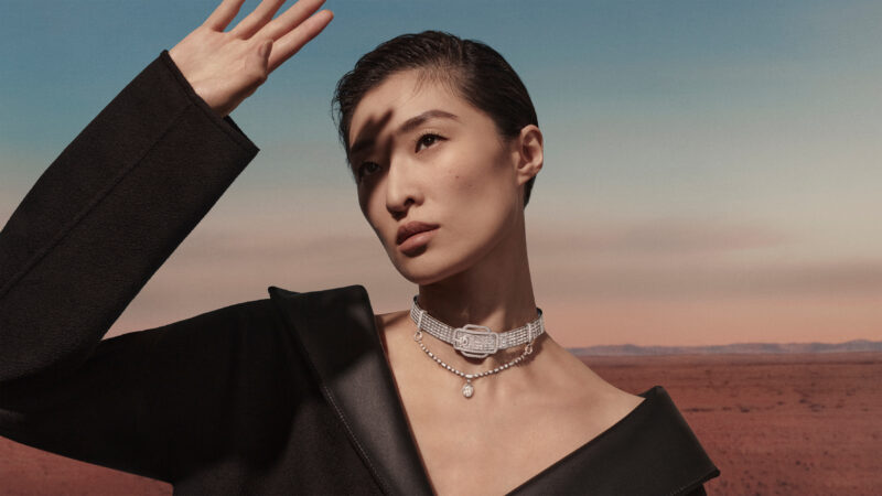 Woman with one arm raised wearing a bright Attelage choker above an open black collar against a low landscape.