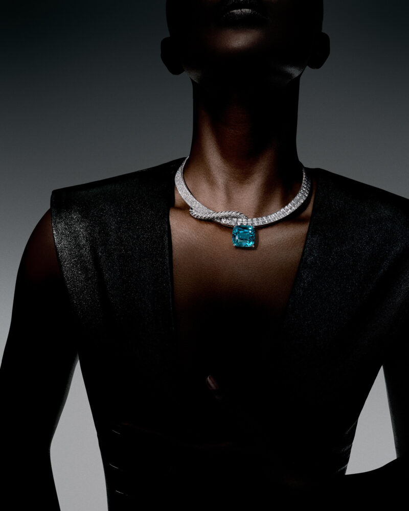 Woman in a dark angular outfit wearing the Fortitude necklace with a large blue centre beneath a bright band.