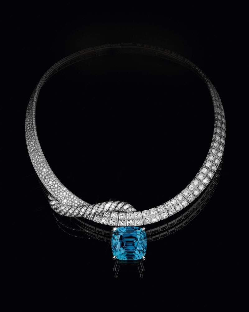 Fortitude necklace on black, a bright curved setting turning towards a large blue cushion-shaped stone.